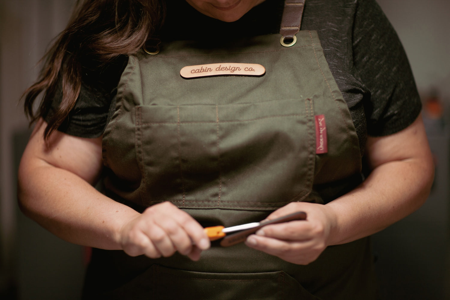 Closeup of female wearing an apron with a leather patch that says Cabin Design Co. She is working on a piece of leather with a tool.