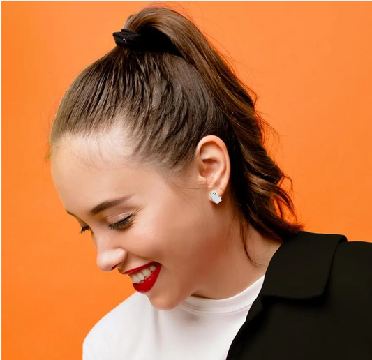 A person wearing a black top and red lipstick, with a smile on their face, showing off their winking ghost stud earrings.