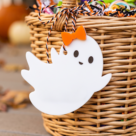 A white ghost-shaped Halloween gift tag with a cute face and an orange bow tie, hanging from a wicker basket filled with treats.