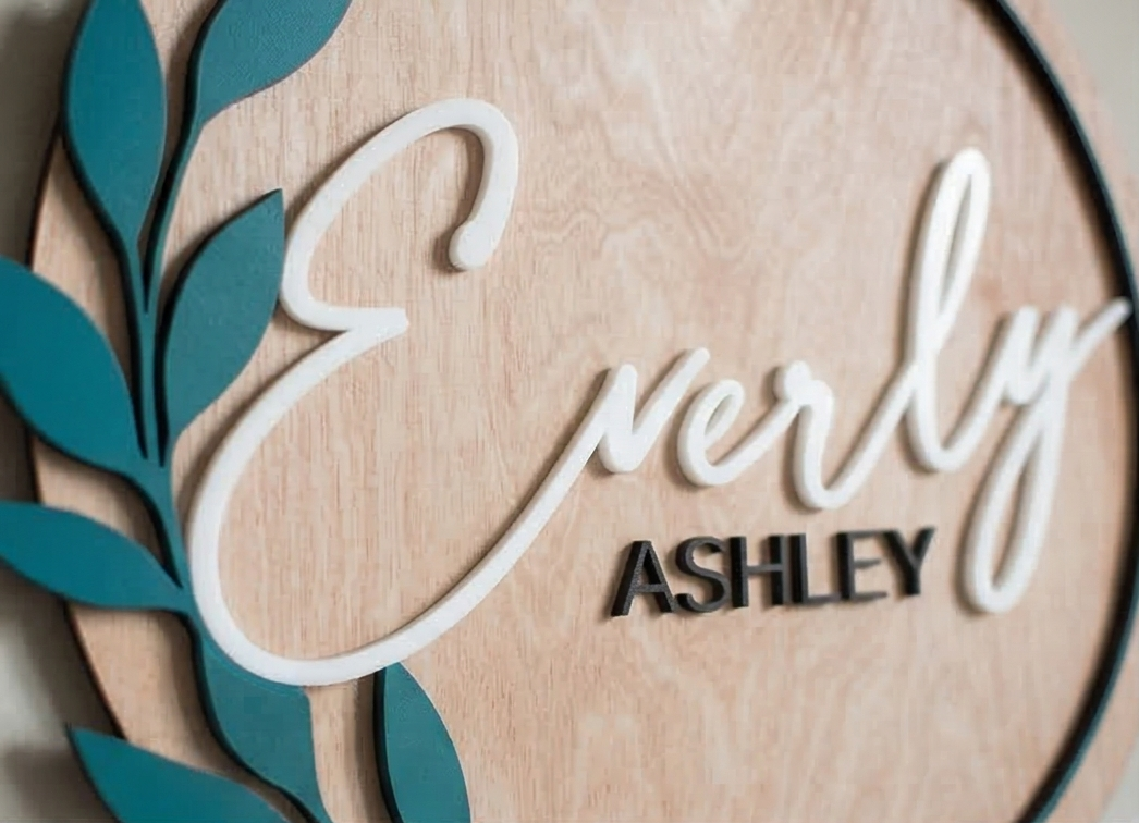 Wooden plaque with 'Everly Ashley' in 3d layer, surrounded by teal leaves.