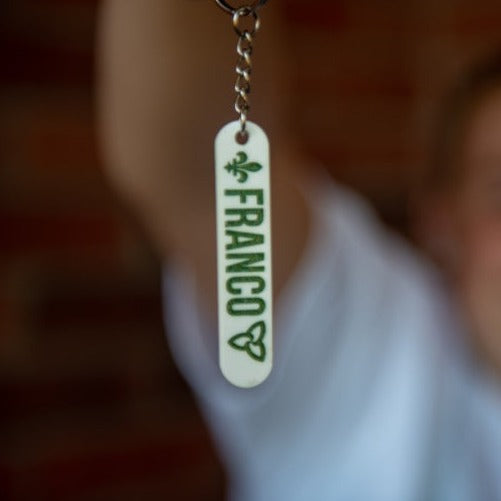 Close-up of a Franco-Ontarian Keychain, a white engraved with "Franco," the trillium, and fleur-de-lys color-filled in green. In the blurred background, a young girl holds the keychain in front of her.