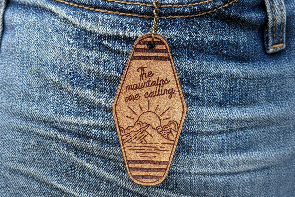 Leather Keychain cut and engraved to look like a vintage motel key with The mountains are calling with an illustration of a sun setting over a mountain engraved on it.