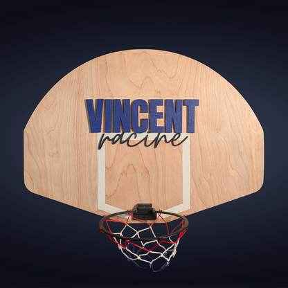 Custom mini basketball hoop with a wooden backboard featuring the name 'Vincent Racine' in layered painted cutouts. The name 'Vincent' is in bold blue 1/4-inch plywood, while 'Racine' is in a cursive style cut from painted 1/8-inch MDF. A 7.5-inch breakaway-style hoop with a red, white, and blue net is attached to the bottom. The piece is displayed on a blue backdrop