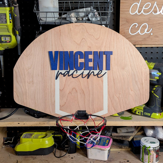 Custom mini basketball hoop with a wooden backboard featuring the name 'Vincent Racine' in layered painted cutouts. The name 'Vincent' is in bold blue 1/4-inch plywood, while 'Racine' is in a cursive style cut from painted 1/8-inch MDF. A 7.5-inch breakaway-style hoop with a red, white, and blue net is attached to the bottom. The piece is displayed in a workshop setting.