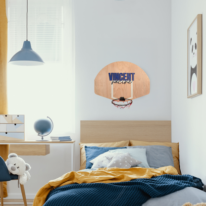 Custom mini basketball hoop with a wooden backboard featuring the name 'Vincent Racine' in layered painted cutouts. The name 'Vincent' is in bold blue 1/4-inch plywood, while 'Racine' is in a cursive style cut from painted 1/8-inch MDF. A 7.5-inch breakaway-style hoop with a red, white, and blue net is attached to the bottom. The piece is displayed in a boy's bedroom above a bed.