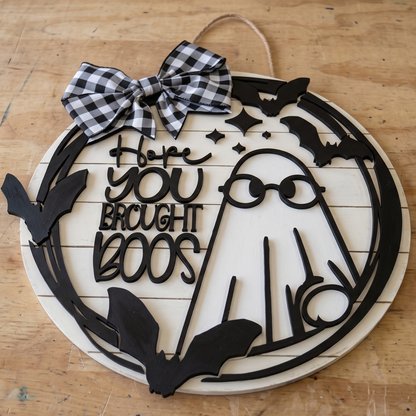 A circular Halloween wooden wall sign with a black laser-cut ghost, bats, stars, and the phrase 'Hope YOU Brought BOOS' in stylized lettering. The sign has a white wood base, a black-and-white gingham bow at the top, and a natural twine hanging loop.