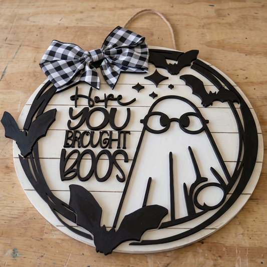 A circular Halloween wooden wall sign with a black laser-cut ghost, bats, stars, and the phrase 'Hope YOU Brought BOOS' in stylized lettering. The sign has a white wood base, a black-and-white gingham bow at the top, and a natural twine hanging loop.