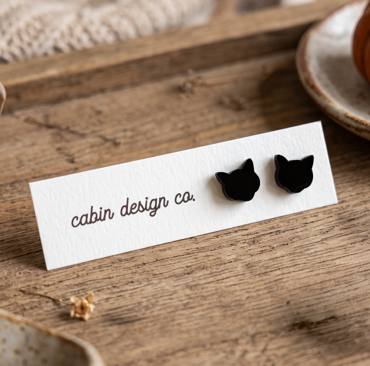 Two small black cat-shaped stud earrings displayed on a white card printed with the brand name 'cabin design co.' The card is placed on a rustic wooden surface with shallow depth of field.