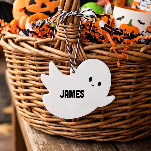 A Halloween-themed wicker basket gift basket filled with candy, a jack-o'-lantern plush, orange crinkle paper, and a white ghost-shaped tag personalized with the name 'JAMES'.