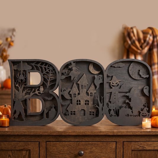 Three large carved wooden letters spelling BOO in a dark charcoal finish, decorated with Halloween motifs including a haunted house, spider, moon, and jack-o'-lanterns, displayed on a wooden cabinet with candles and pumpkins in a warm indoor setting.