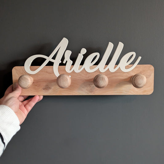 Personalized wooden towel rack with the name "Arielle" in white script on a natural wood base with four rounded pegs, handmade from layered plywood.