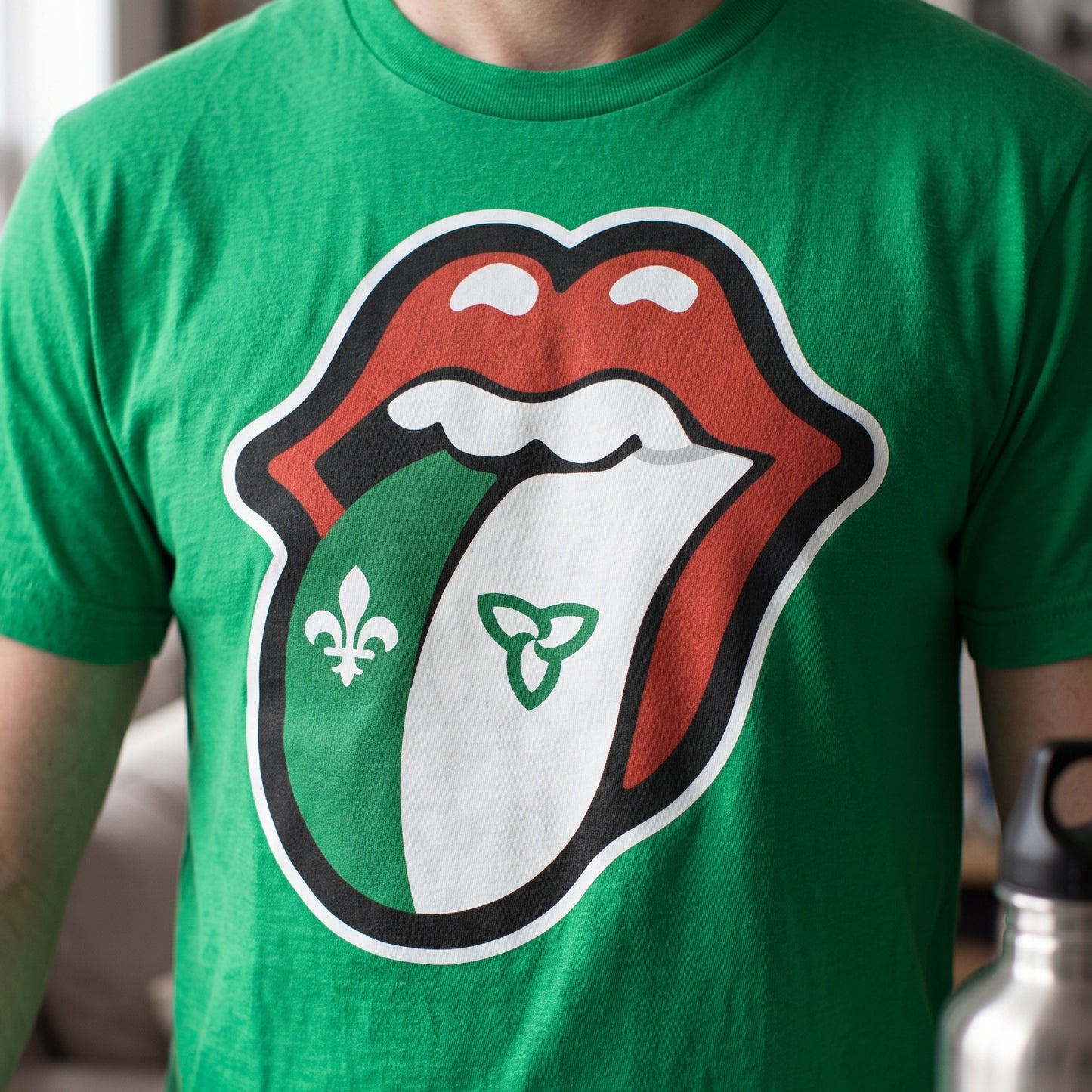 A green short-sleeve t-shirt featuring a large graphic of the Rolling Stones tongue logo, with the left side of the tongue colored green and decorated with a fleur-de-lis, and the right side colored white with a green Celtic triquetra symbol.