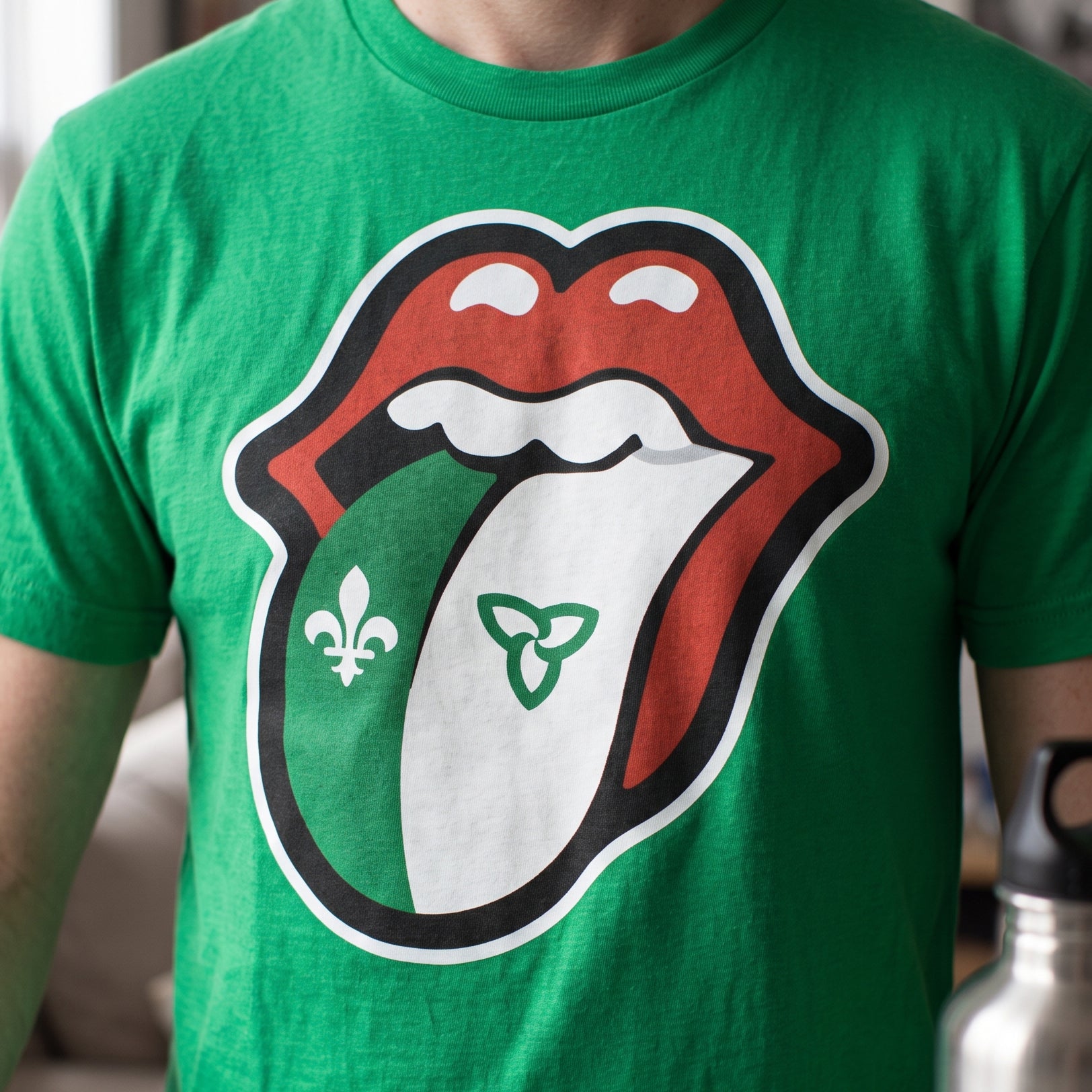 A green short-sleeve t-shirt featuring a large graphic of the Rolling Stones tongue logo, with the left side of the tongue colored green and decorated with a fleur-de-lis, and the right side colored white with a green Celtic triquetra symbol.
