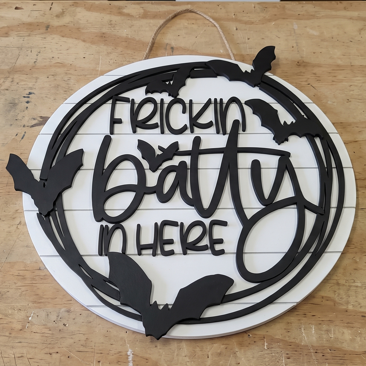 A circular white wooden hanging sign with black cutout lettering that reads 'Frickin Batty In Here' and black bat silhouettes around the border, displayed on a light wood surface.