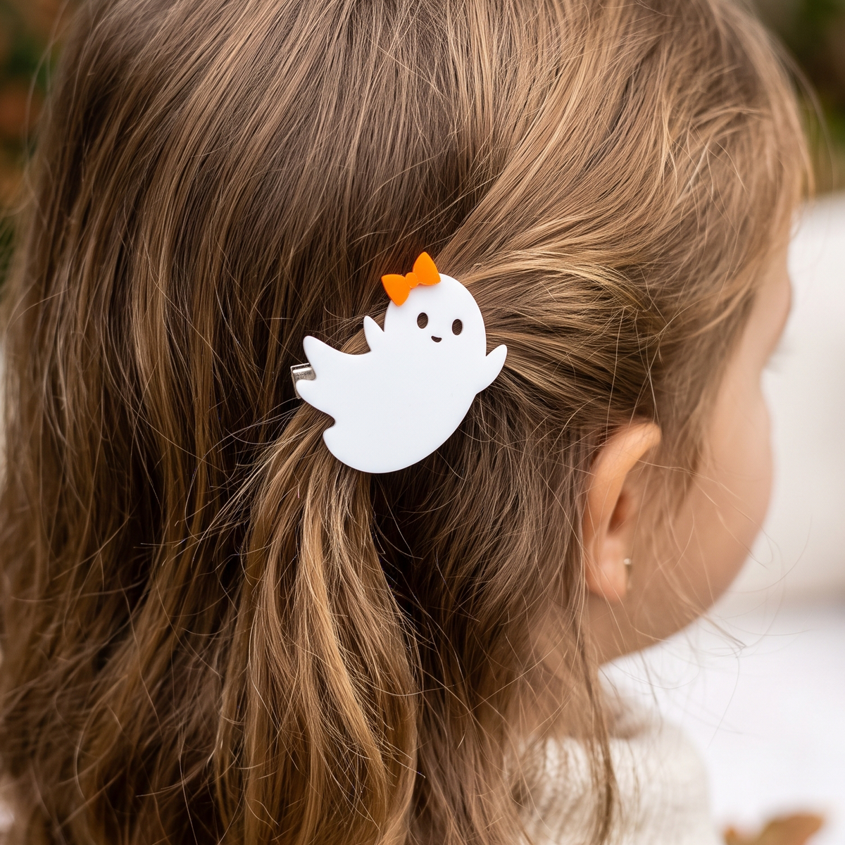 A white ghost-shaped hair clip with a small orange bow, shown clipped to a child's light brown hair in a close-up side profile view.