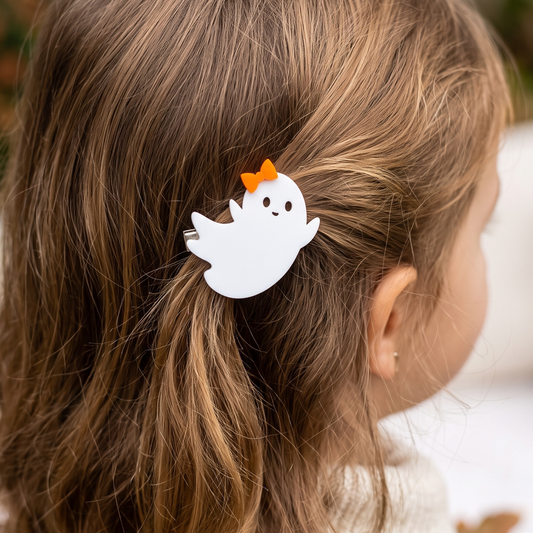 A white ghost-shaped hair clip with a small orange bow, shown clipped to a child's light brown hair in a close-up side profile view.