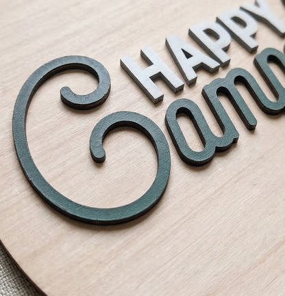 Wooden cutout letters spelling 'Happy Camper' on a wooden background
