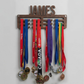 Medal holder with medals and ribbons hanging on a white wall, personalized with 'JAMES'.