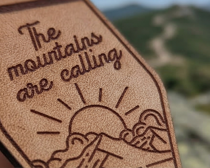 Leather keychain with 'The mountains are calling' text and mountain illustration, blurred mountain landscape in the background.