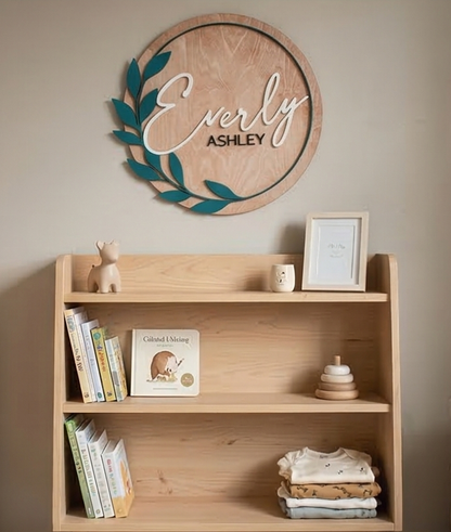 Wooden shelf with books, toys, and a framed picture below a decorative wall sign with 'Everly Ashley' on a beige wall.