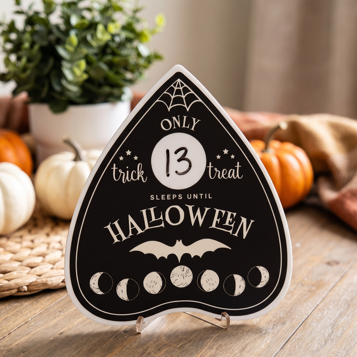 A Halloween-themed ouija board shaped sign on a small stand, reading 'ONLY 13 TRICK treats SLEEPS UNTIL HALLOWEEN,' styled on a wooden surface with pumpkins, a plant, and a draped fabric in the background.