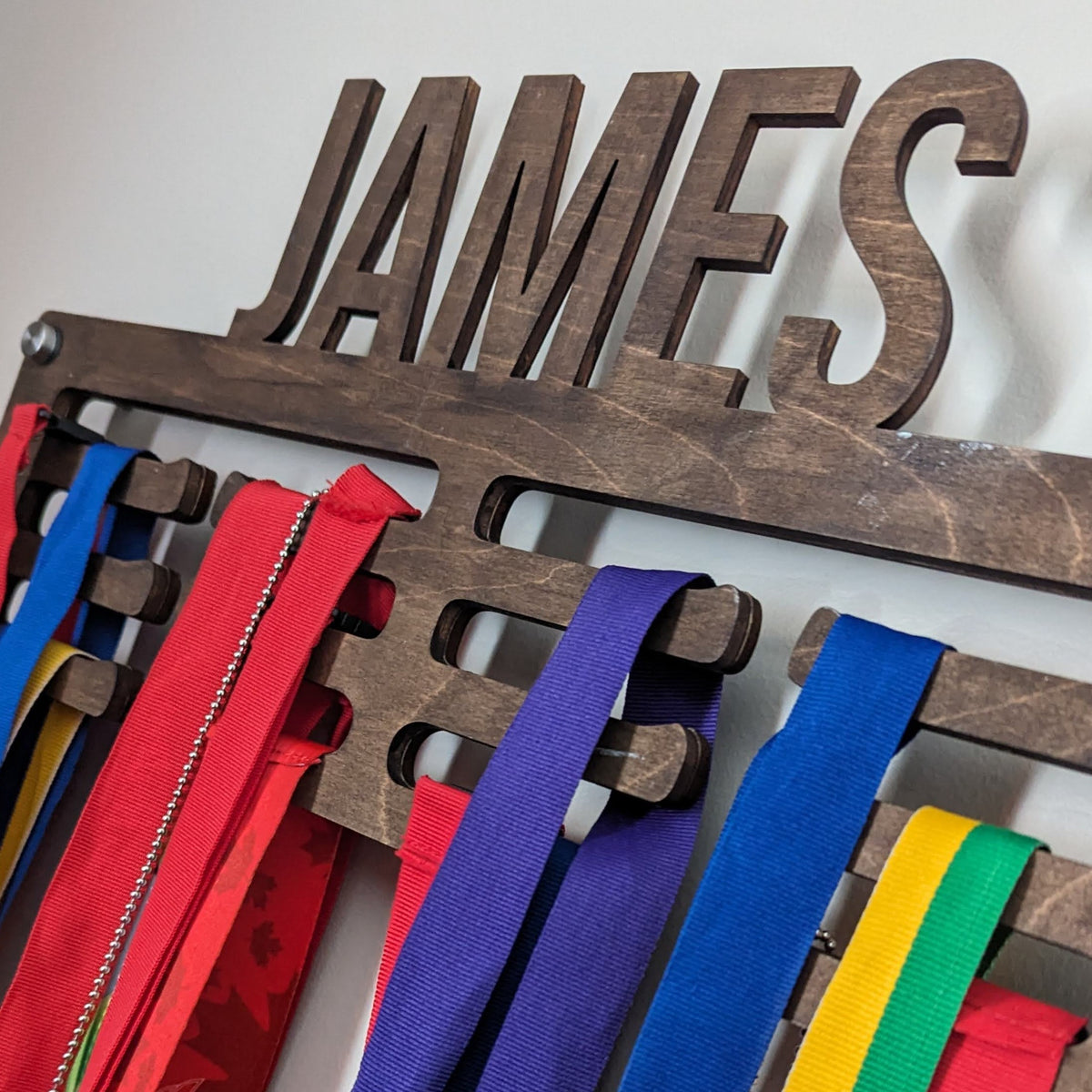 Close-up of a customized with the name James wooden medal holder mounted on a wall, with red green, yellow and blue ribbons hanging from it