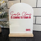 A white acrylic countdown sign with a wooden base reads “Santa Claus is coming to town in ___ days” in red lettering. The sign has a modern arched top and sits on a black countertop beside a snow-covered Christmas tree branch.