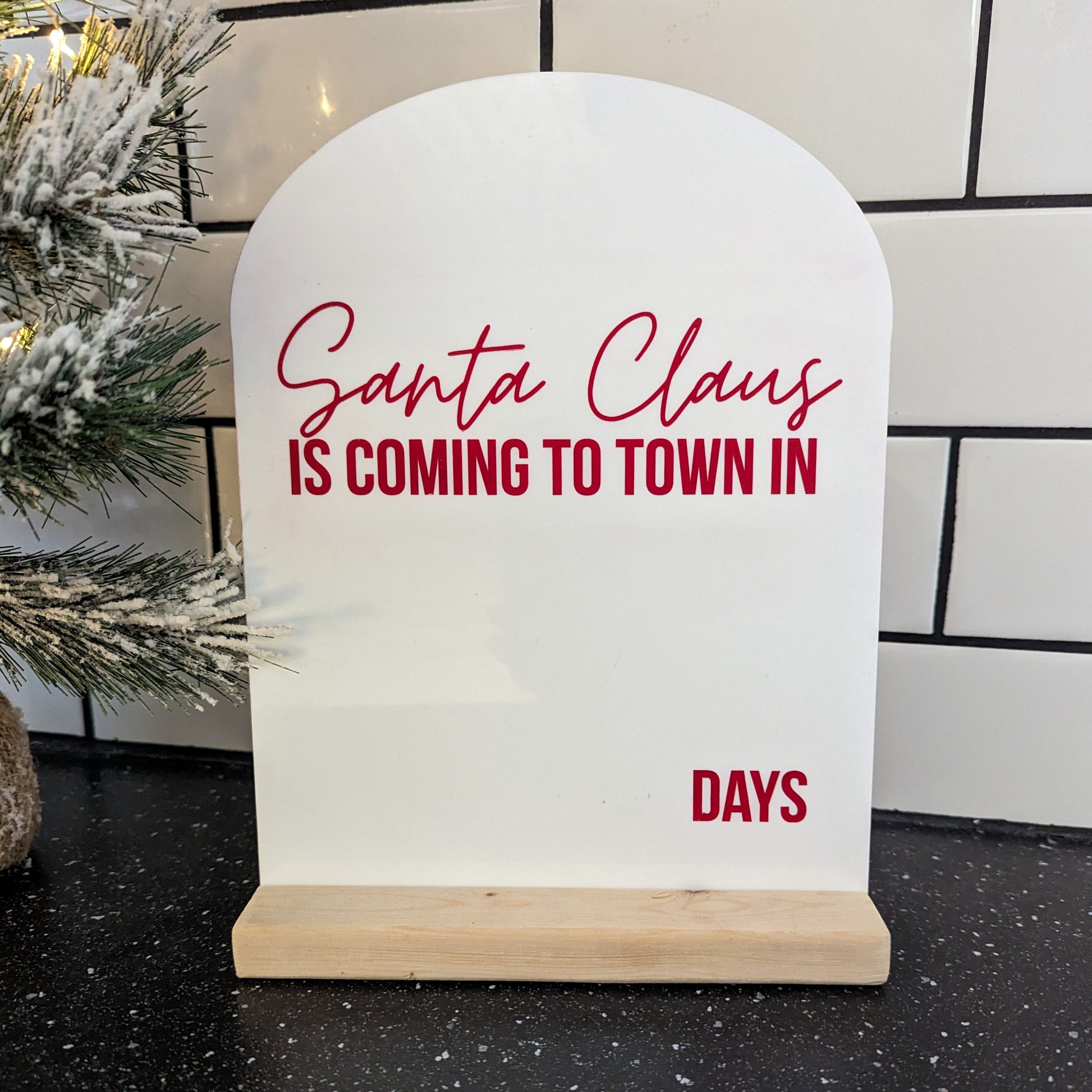 A white acrylic countdown sign with a wooden base reads “Santa Claus is coming to town in ___ days” in red lettering. The sign has a modern arched top and sits on a black countertop beside a snow-covered Christmas tree branch.